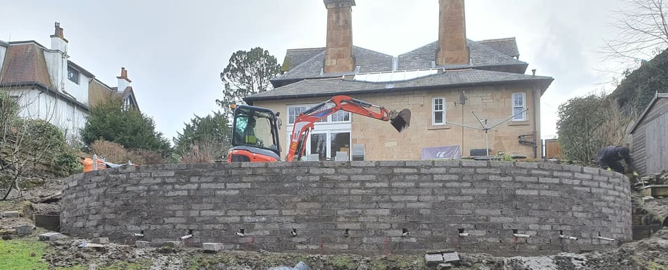 Curved wall of grey concrete blockwork with white weep pipes near its base, a mini digger and the house behind.