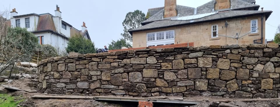 Curved garden retaining wall faced in buff, brown and grey random rubble sandstone.