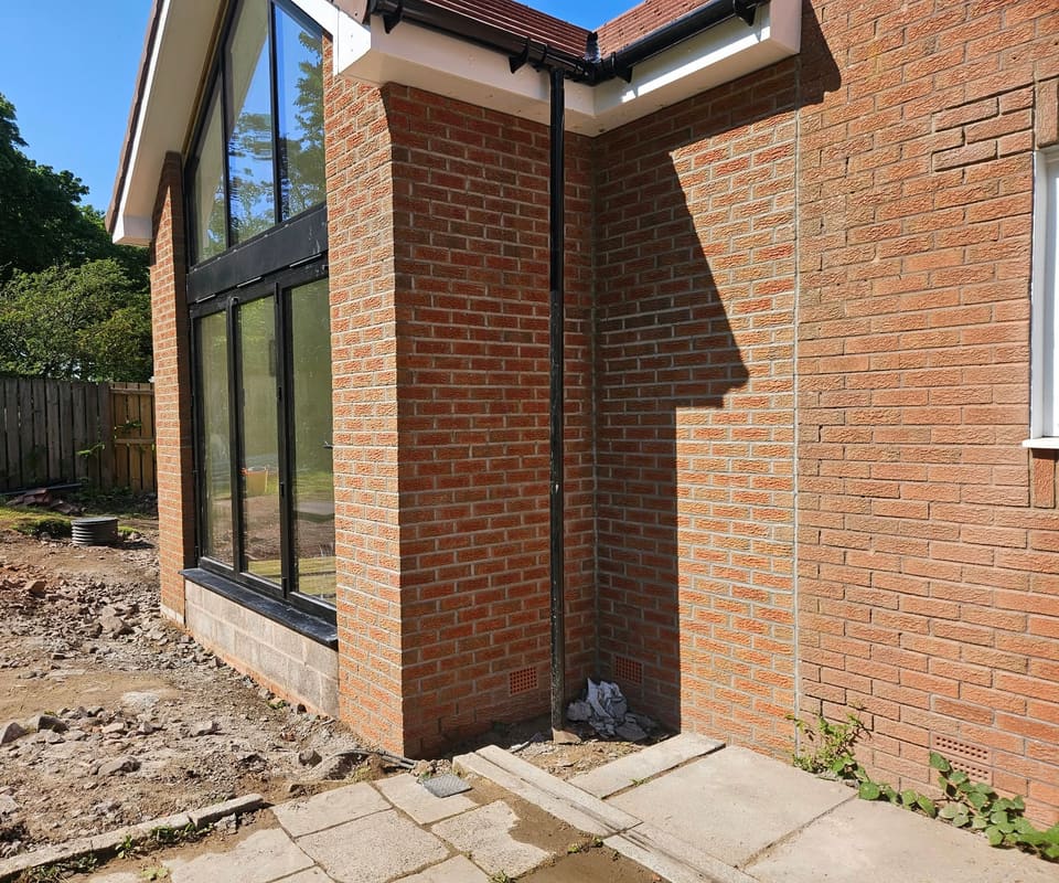 Brick pier beside black-framed glazing and an internal corner with a downpipe. A straight vertical joint runs up the wall to the right.