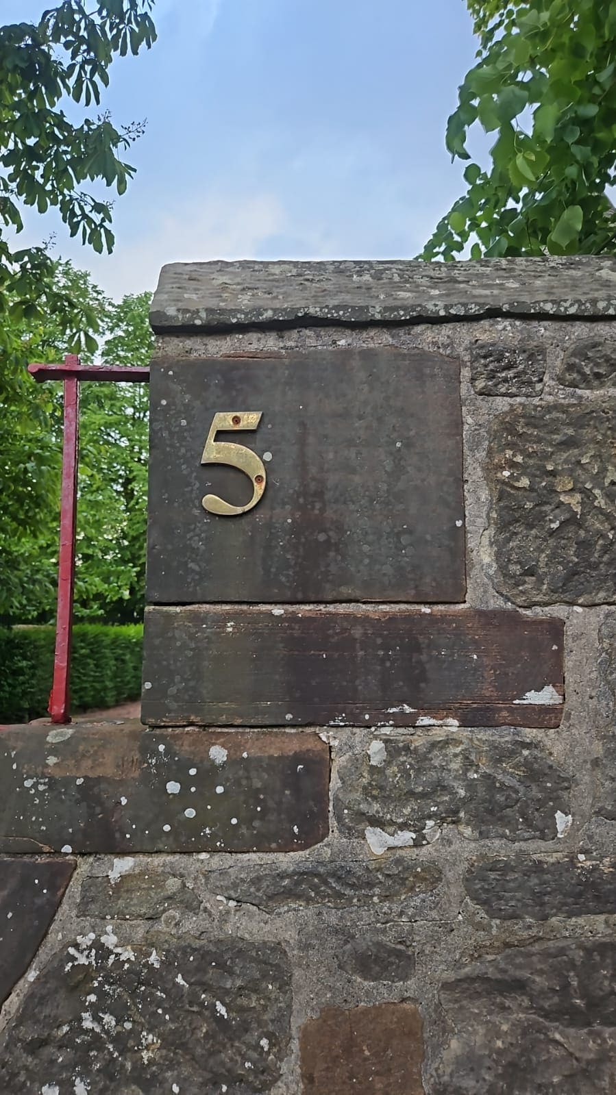 Stone gate pier with a plain dark panel carrying a brass number 5, a blank stone band below and red-painted gate ironwork to the left.