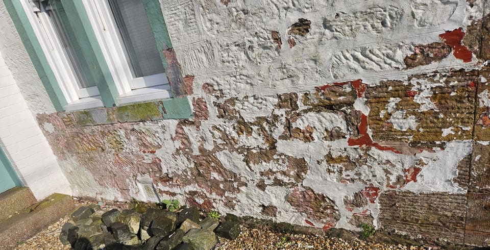 Close view of white paint peeling off tooled sandstone blocks, with red and pink coats beneath and a green-painted window margin.