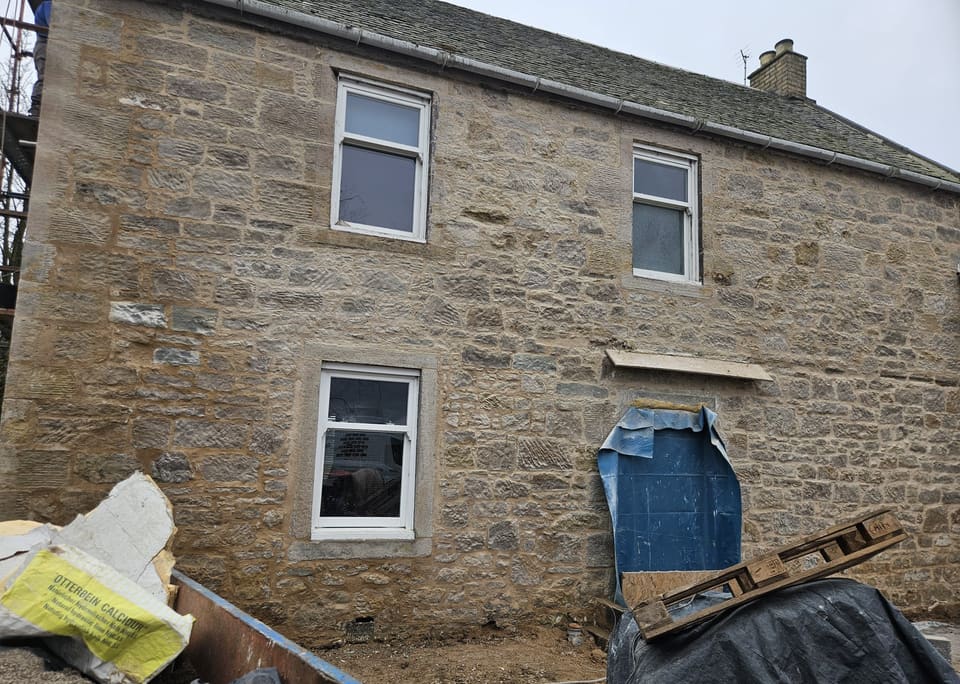 Two-storey rubble wall repointed in buff mortar, windows uncovered, doorway sheeted. An Otterbein Calcidur NHL 3.5 lime bag lies in a skip.