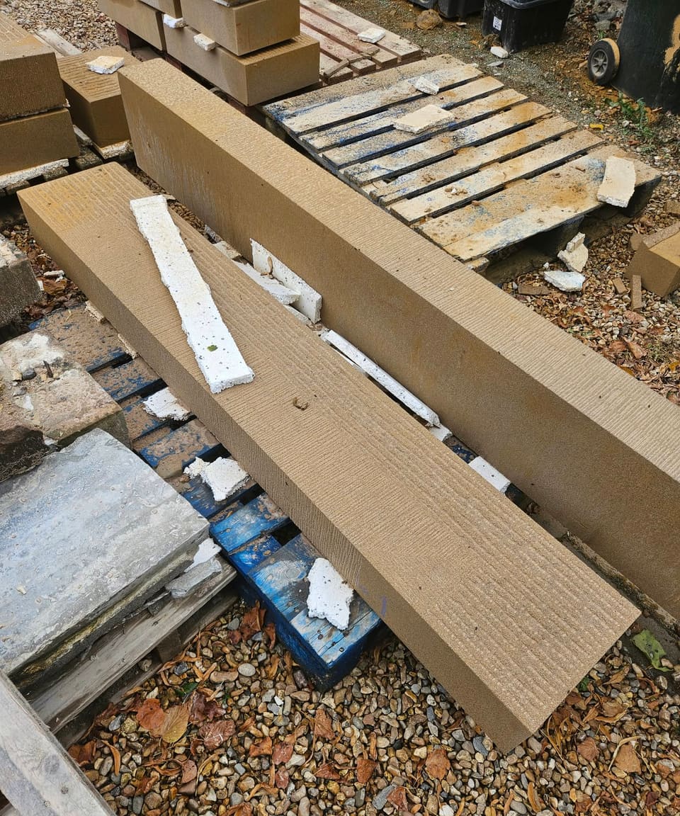 Two long new sandstone lintels with broached faces lying across blue pallets on gravel, smaller new blocks stacked behind.