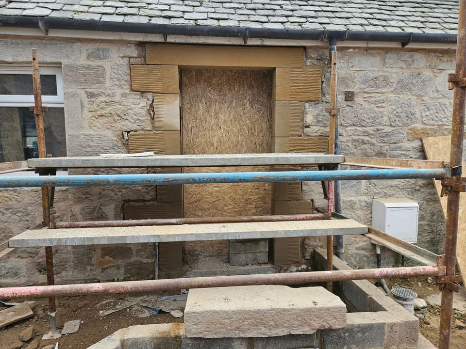 Window opening taken down and rebuilt as a doorway, with new sandstone rybats and lintel, behind a scaffold.