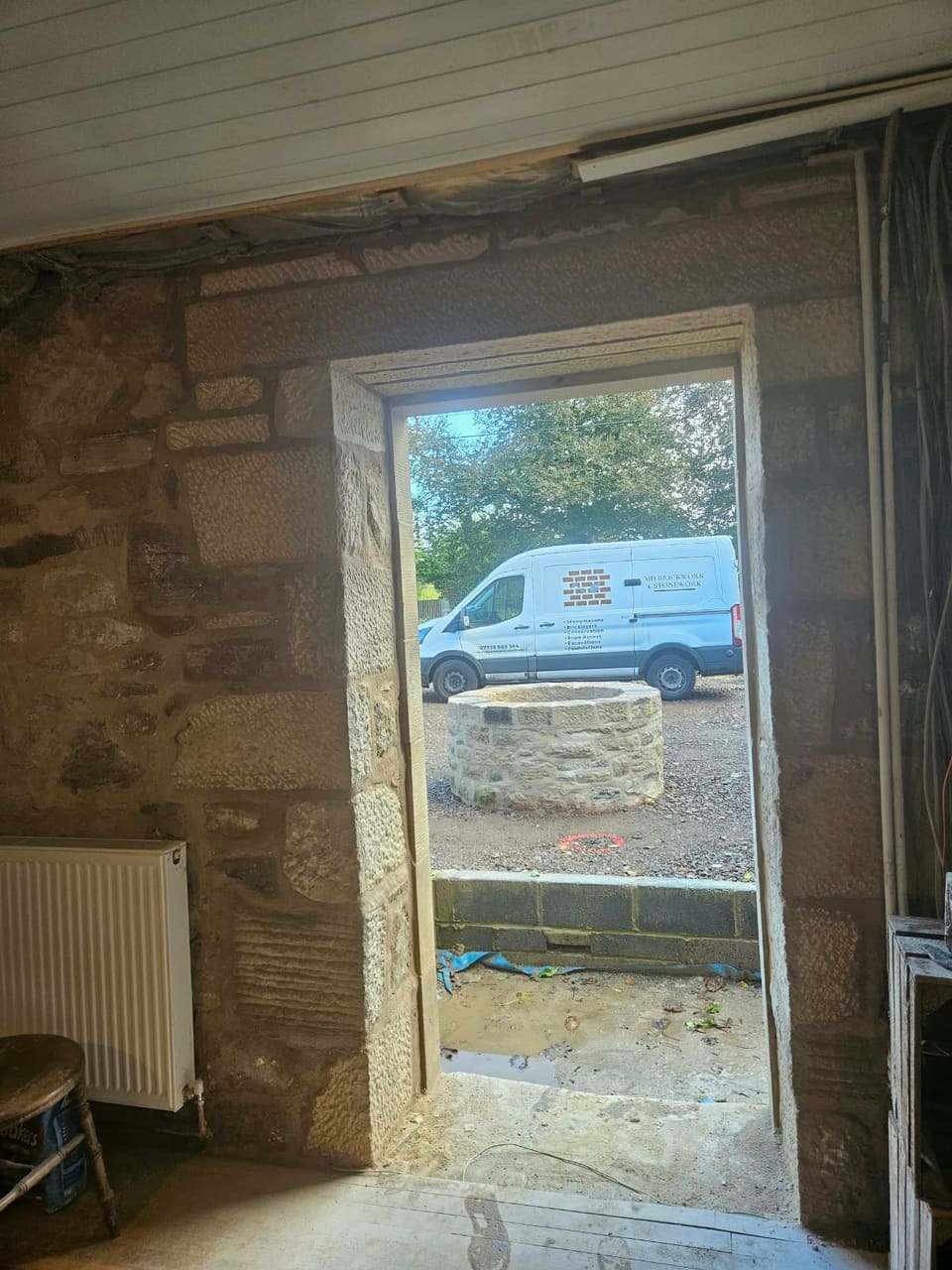 View from inside out through the new doorway to the rebuilt round stone well, with a white MD Brickwork & Stonework van parked beyond.