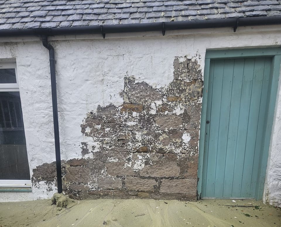 White-painted stone wall with a large patch stripped back to brown sandstone, a white window to the left and the blue-green door to the right.