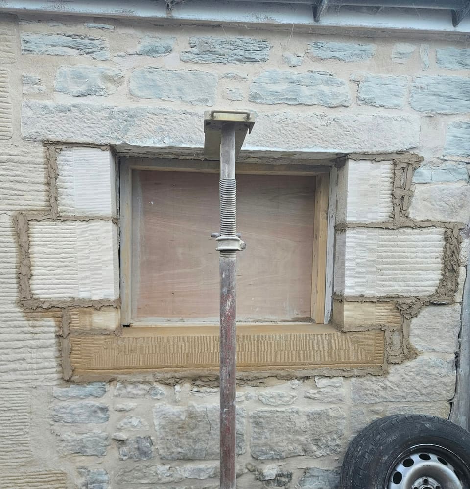 Small window opening boarded with plywood, new sandstone rybats, sill and lintel bedded in fresh mortar, an adjustable prop in front.