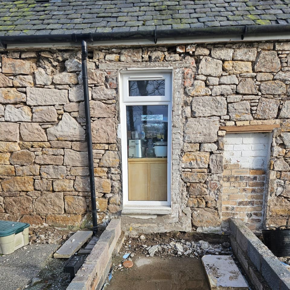 Tall white window with a top light in a rubble stone wall, a downpipe to its left.