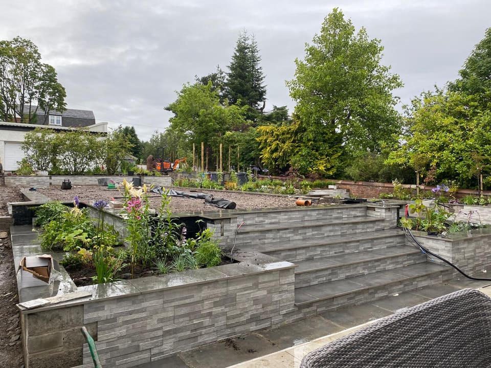Wide clad steps rising between raised planters with flowering plants, slab copings on the walls, the upper level still to be finished.