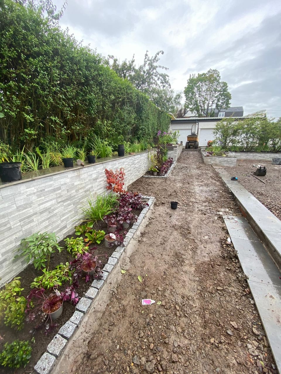Long clad wall down the side of the garden with a planted bed and granite sett edging, a path being formed alongside.