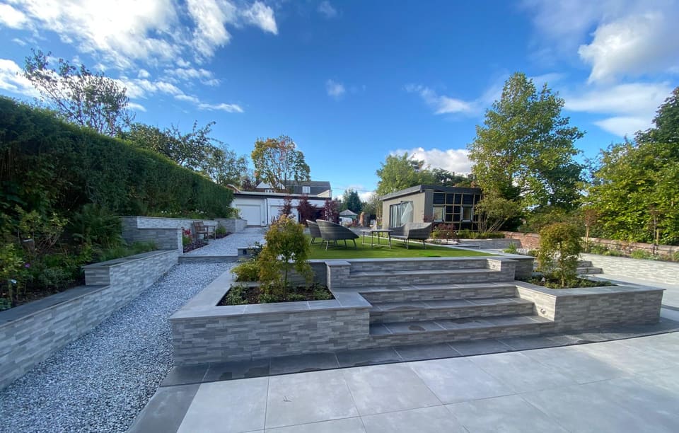 Two-level garden with a grey paved patio, tiled steps between raised planters, clad walls and a lawn terrace.