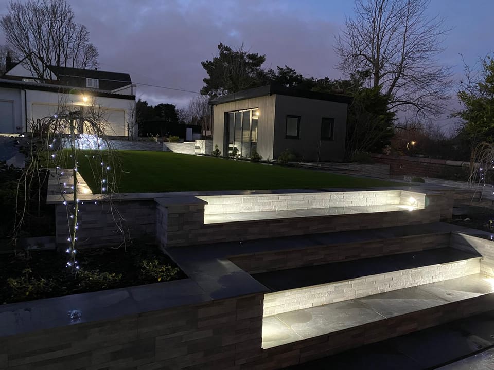 Lit steps between planters at dusk, with the lawn and a garden room behind.