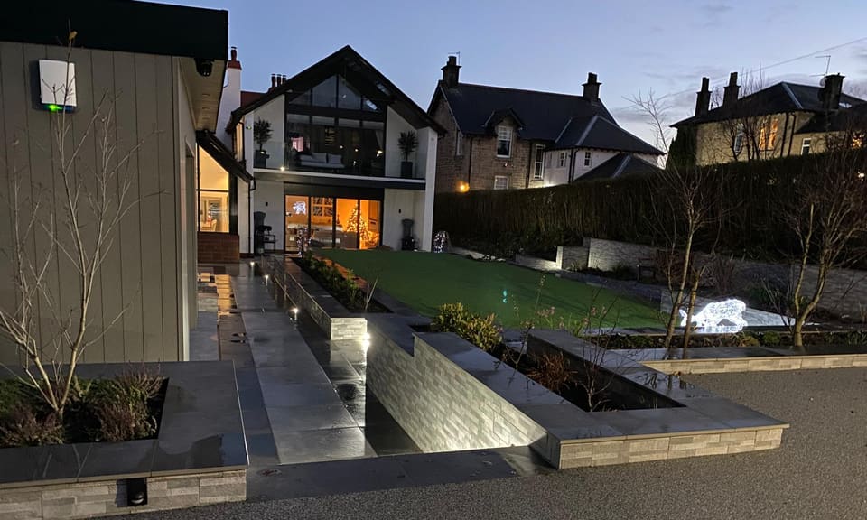Lit clad walls and dark paving leading towards the house at dusk.