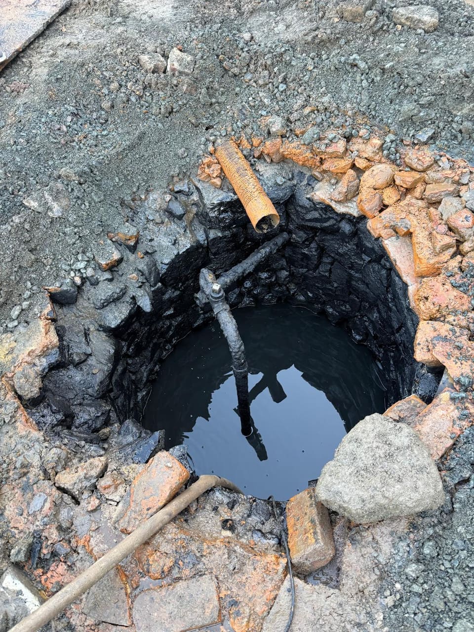 Well opening with broken red brick round the rim, black-coated stone lining, an old iron pipe with a valve and a hose running into water.
