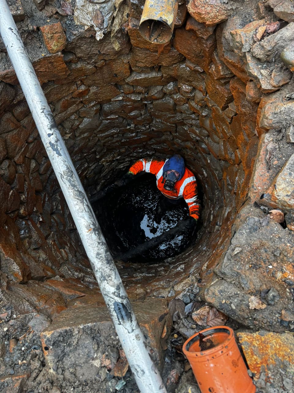 View down the shaft to a worker in orange hi-vis, blue hood and respirator standing in black sludge, a metal pole running down the shaft.