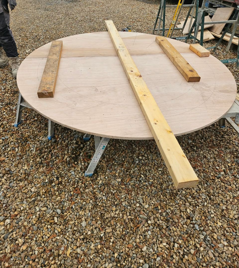 Large plywood disc made from two joined sheets, marked with faint pencil circles, on low work platforms on gravel, timber battens on top.