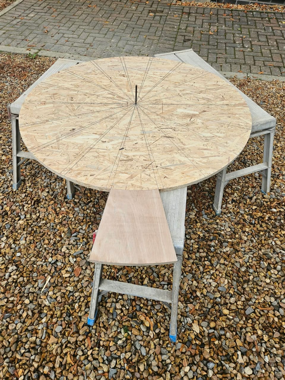Circular OSB board on three low work platforms, pencil lines radiating from a centre pin, a tapered plywood pattern laid at its near edge.