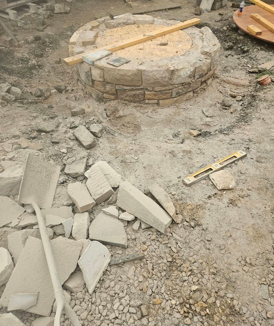 First courses of a round stone well head above ground, with an OSB disc sitting in the opening.