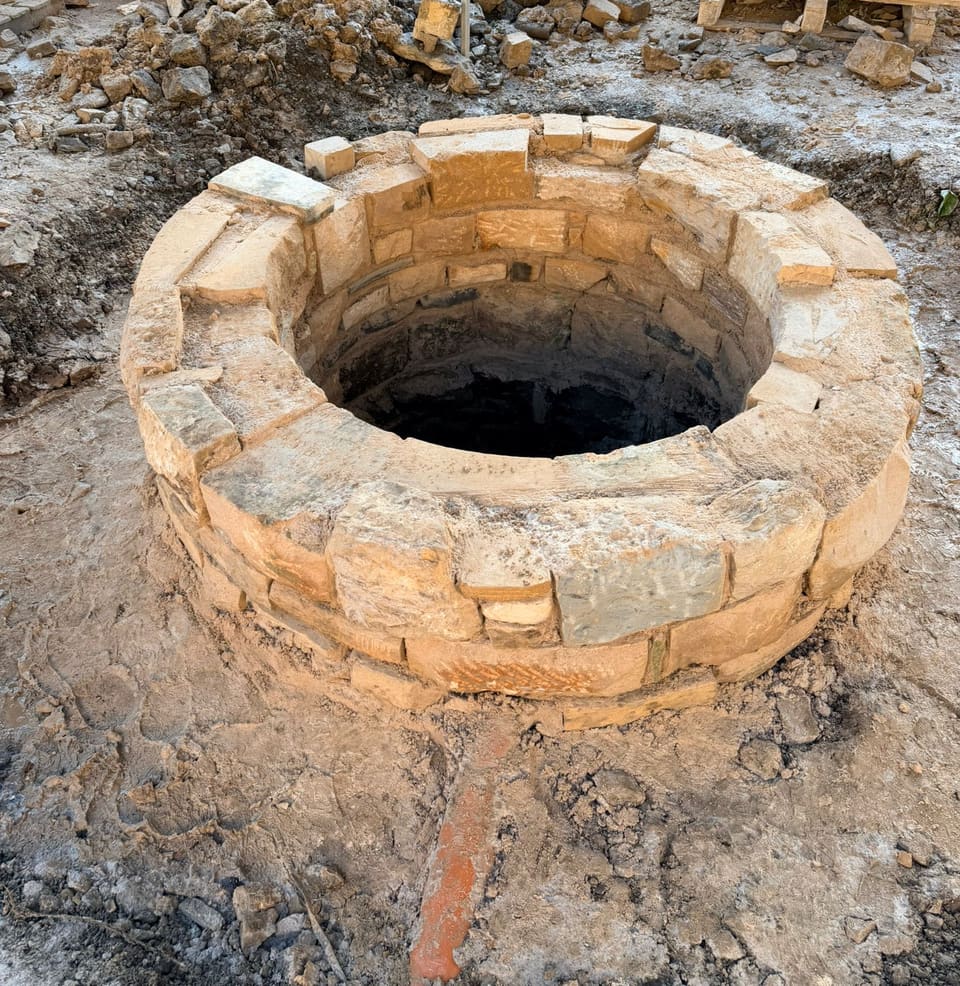 Open round well head a few courses high in buff stone, top course part laid, new stonework inside above the dark shaft, orange pipe in front.