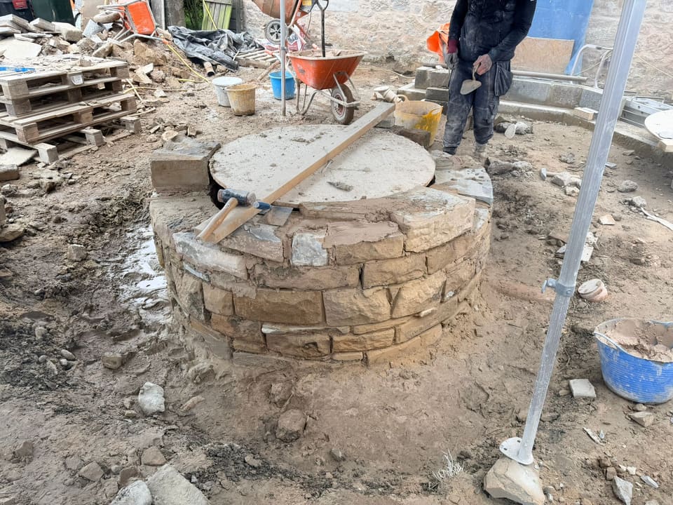 Round stone well head several courses high, fresh mortar on top, a dusty disc over the opening with a mallet and trowel on it, mason behind.