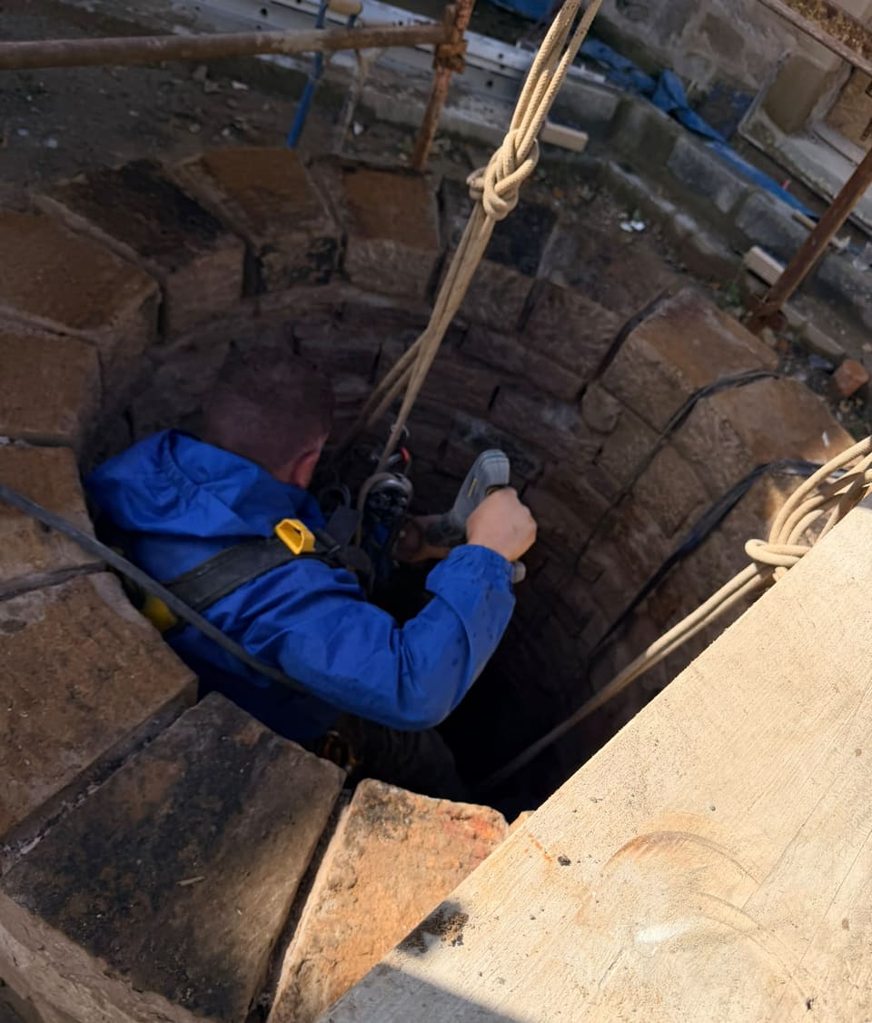 From the scaffold: a worker in blue waterproofs and harness on ropes inside the top of the well, holding a cordless tool to the inner face.