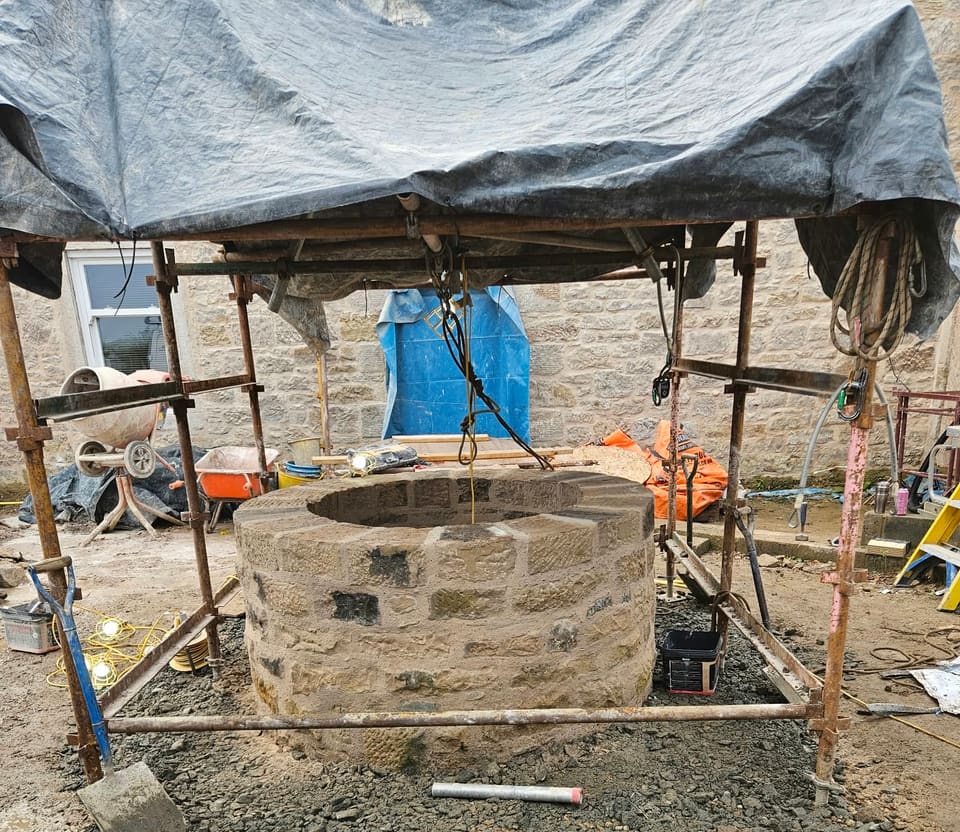 Round stone well head, flush pointed, under a scaffold frame roofed with a black tarpaulin, ropes hanging from it, stone house wall behind.