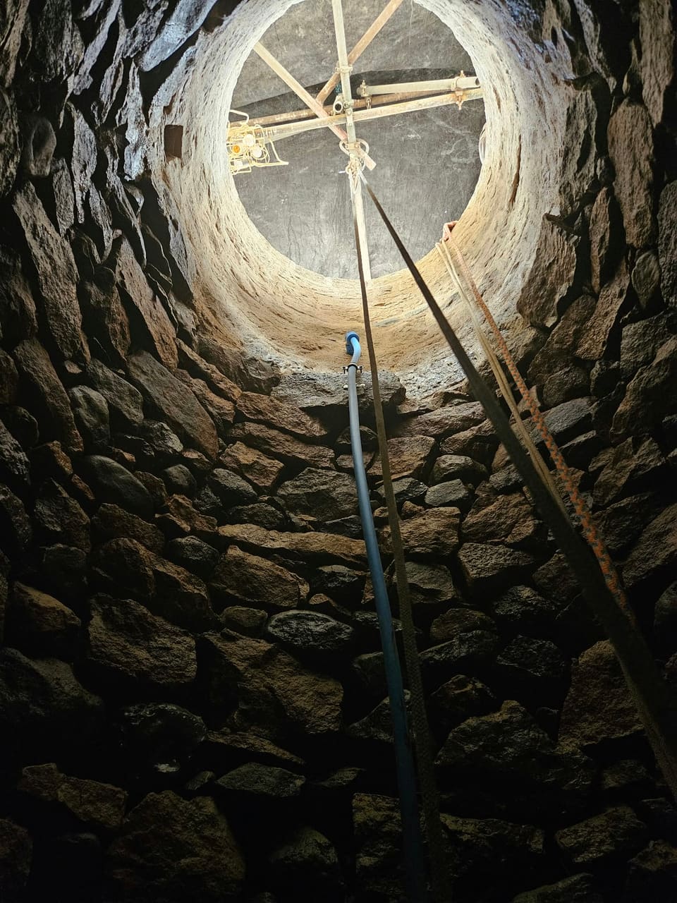 Looking up the well from the bottom: dark original rubble lining, the new stonework lit near the top, and a frame of poles with work lights over the opening.