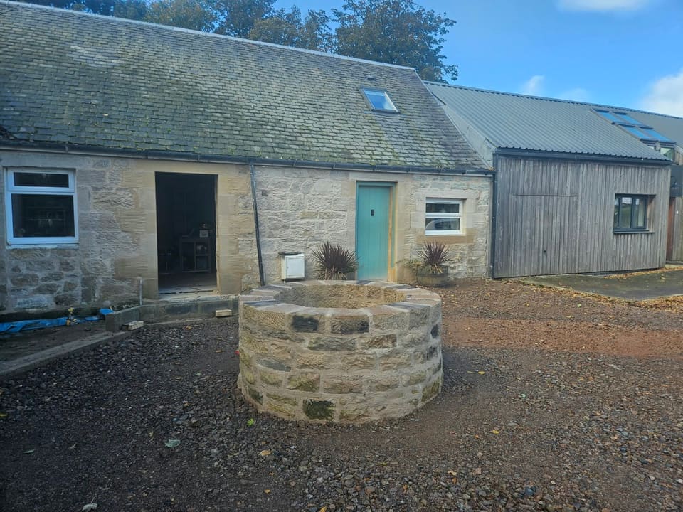 Finished round stone well head in a gravelled garden in front of a low sandstone building.