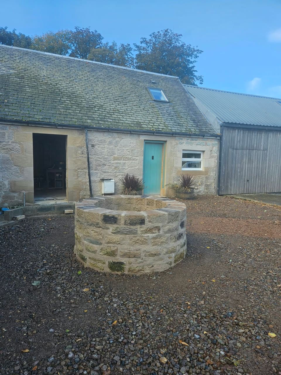 Finished round stone well head with a ring of coping stones, standing in a gravelled garden in front of a low stone building.