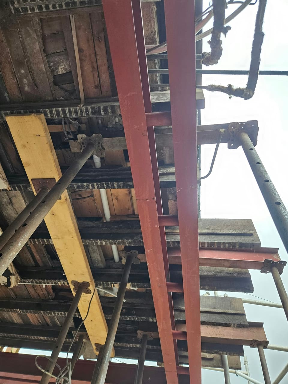 Looking up at red steel beams carrying the floor joists, with adjustable props and needles still in.